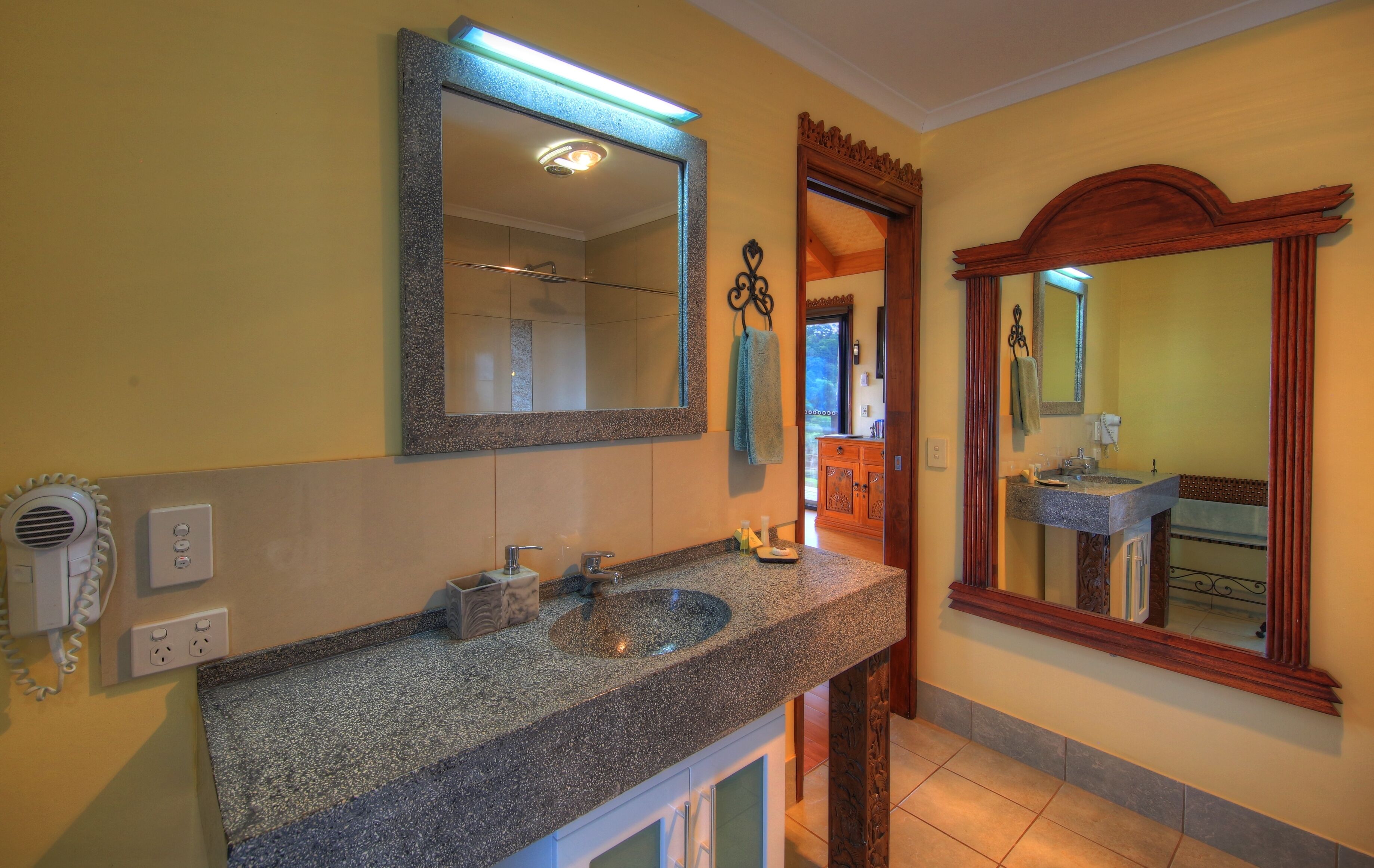 Un baño con lavabo de encimera de granito, un espejo grande y una ducha visible al fondo.