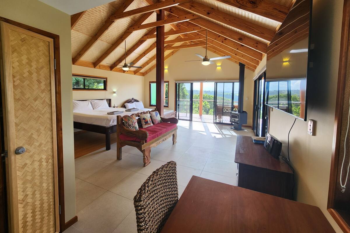 A spacious room with a high ceiling, wooden beams, a bed, a sofa, a dining table, and a TV.