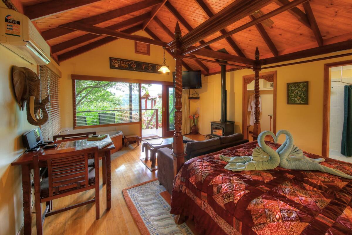 A cozy room with a wooden ceiling, a large bed with a red quilt, a wooden chair, a desk with a computer, and a fireplace.
