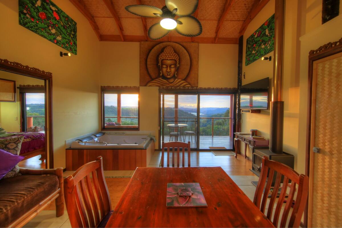 A dining area with a wooden table, chairs, a Buddha statue, a hot tub, and a view of the mountains.