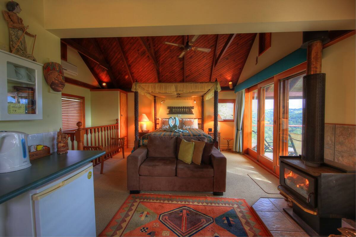 A cozy living room with a wooden ceiling, a sofa, a coffee table, a fireplace, and a view through large windows.