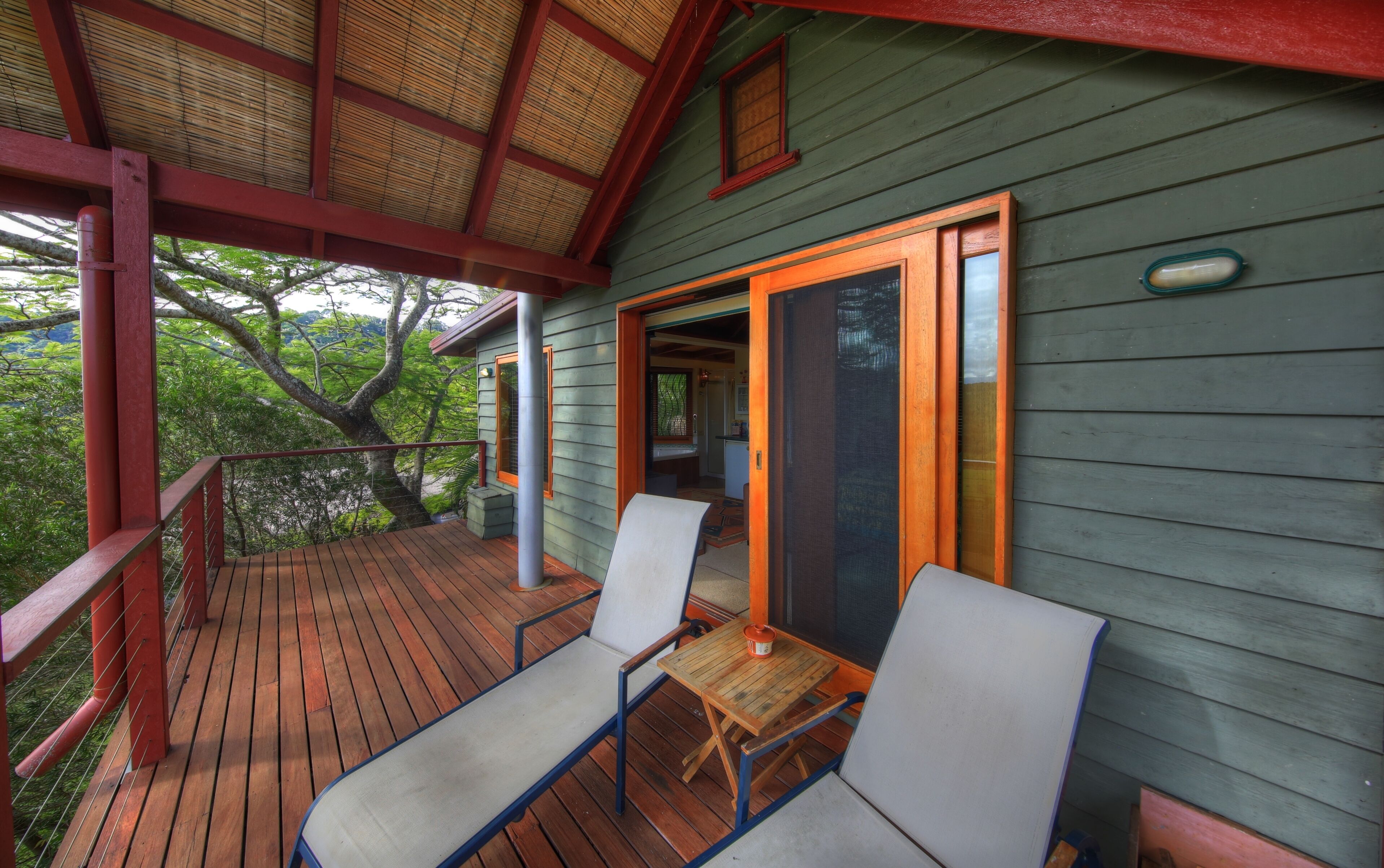 Een houten terras met twee stoelen en een tafel, een overdekte veranda met een rode balustrade, en een groen gebouw met een schuifpui.