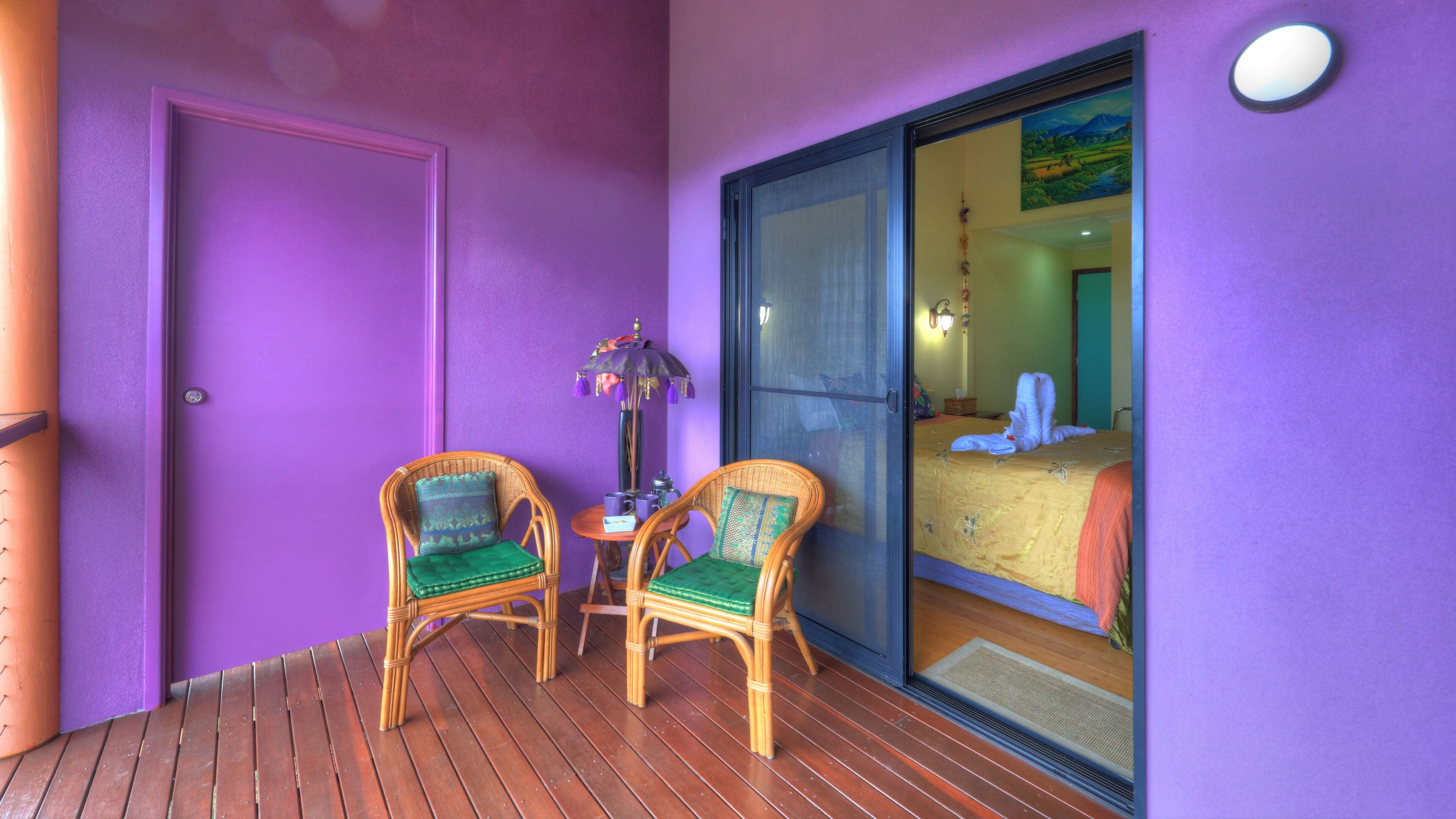 A balcony with two wicker chairs, a small table with a vase, and a sliding glass door leading to a bedroom with a bed and a painting.