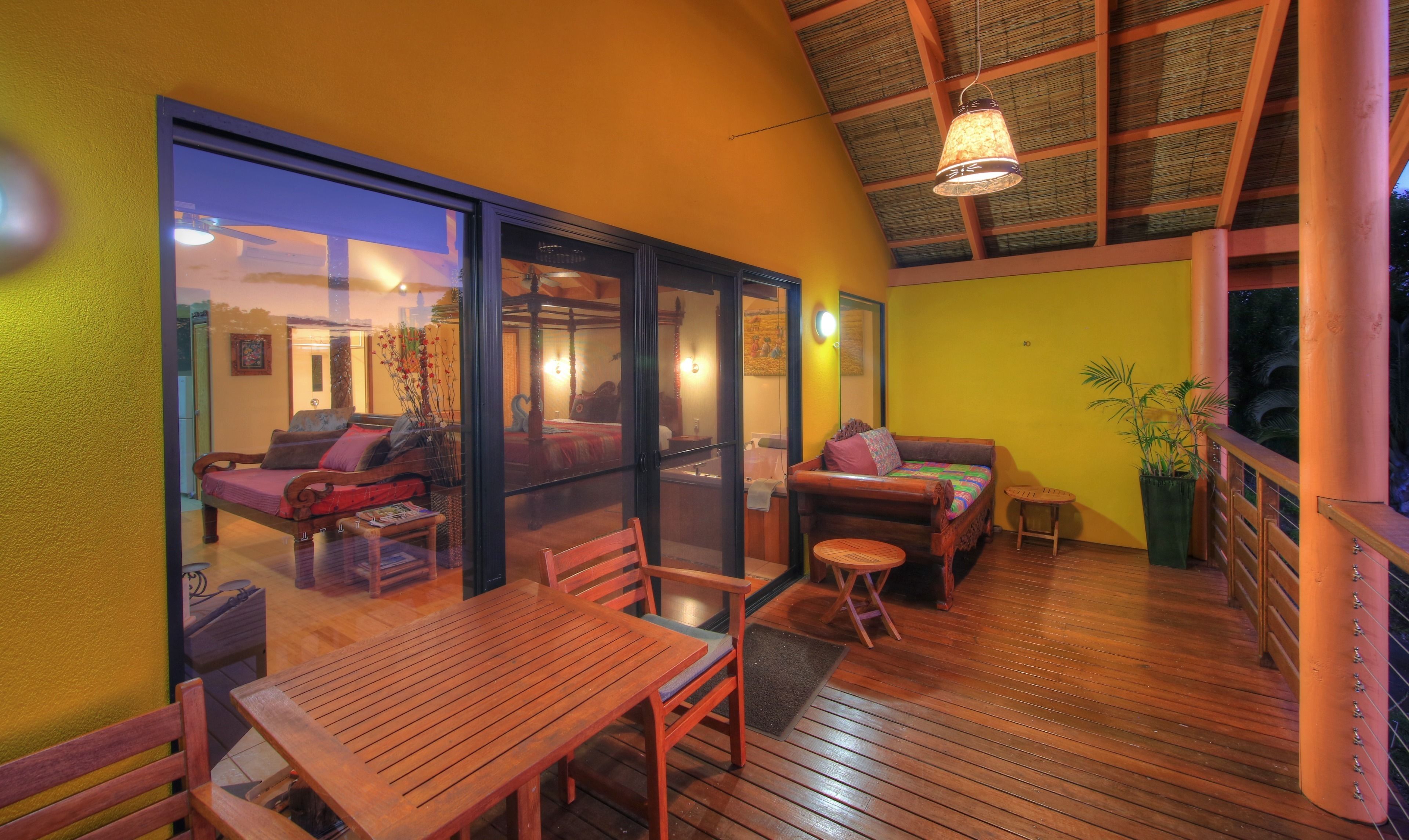 Un intérieur chaleureux avec une terrasse en bois, un coin repas et une vue, à travers une porte vitrée, sur une pièce comprenant un lit et un canapé.