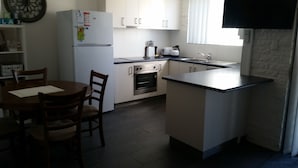 A compact kitchen with a refrigerator, oven, sink, and a dining area with chairs. - Apollo Holiday Units (Narooma)