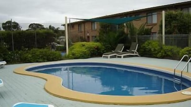 A swimming pool with a curved edge, surrounded by a tiled deck. There are lounge chairs and a shaded area. - Apollo Holiday Units (Narooma)
