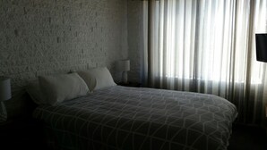 A hotel room with a brick-patterned wall, a large bed with a geometric-patterned bedspread, two bedside lamps, and sheer curtains allowing natural light. - Apollo Holiday Units (Narooma)