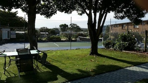 A tennis court with a fence, trees, and a table with chairs in the foreground. - Apollo Holiday Units (Narooma)