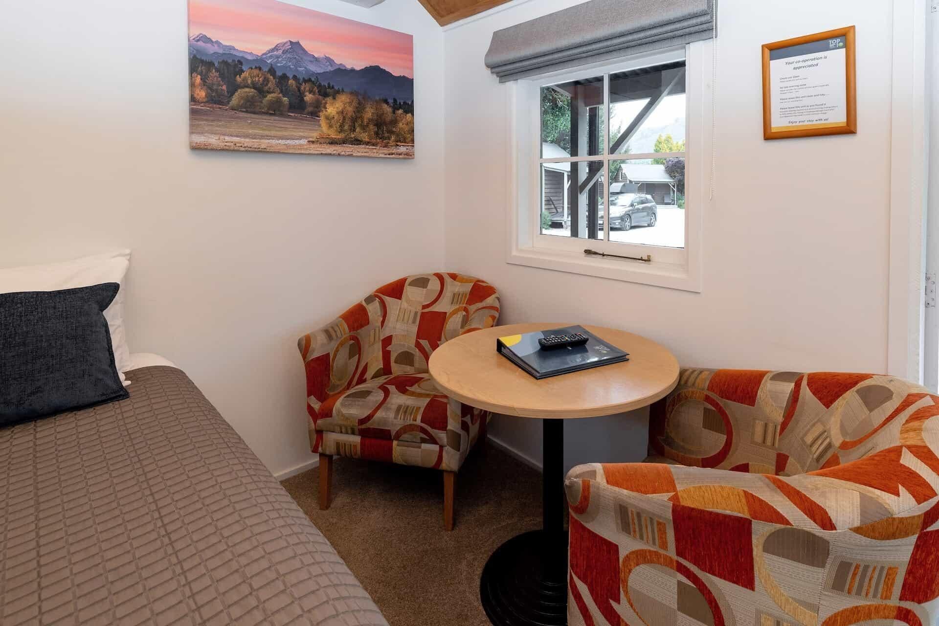 A small, cozy room with a bed, two patterned armchairs, a small round table with a remote control, and a window with a view of a car and trees.