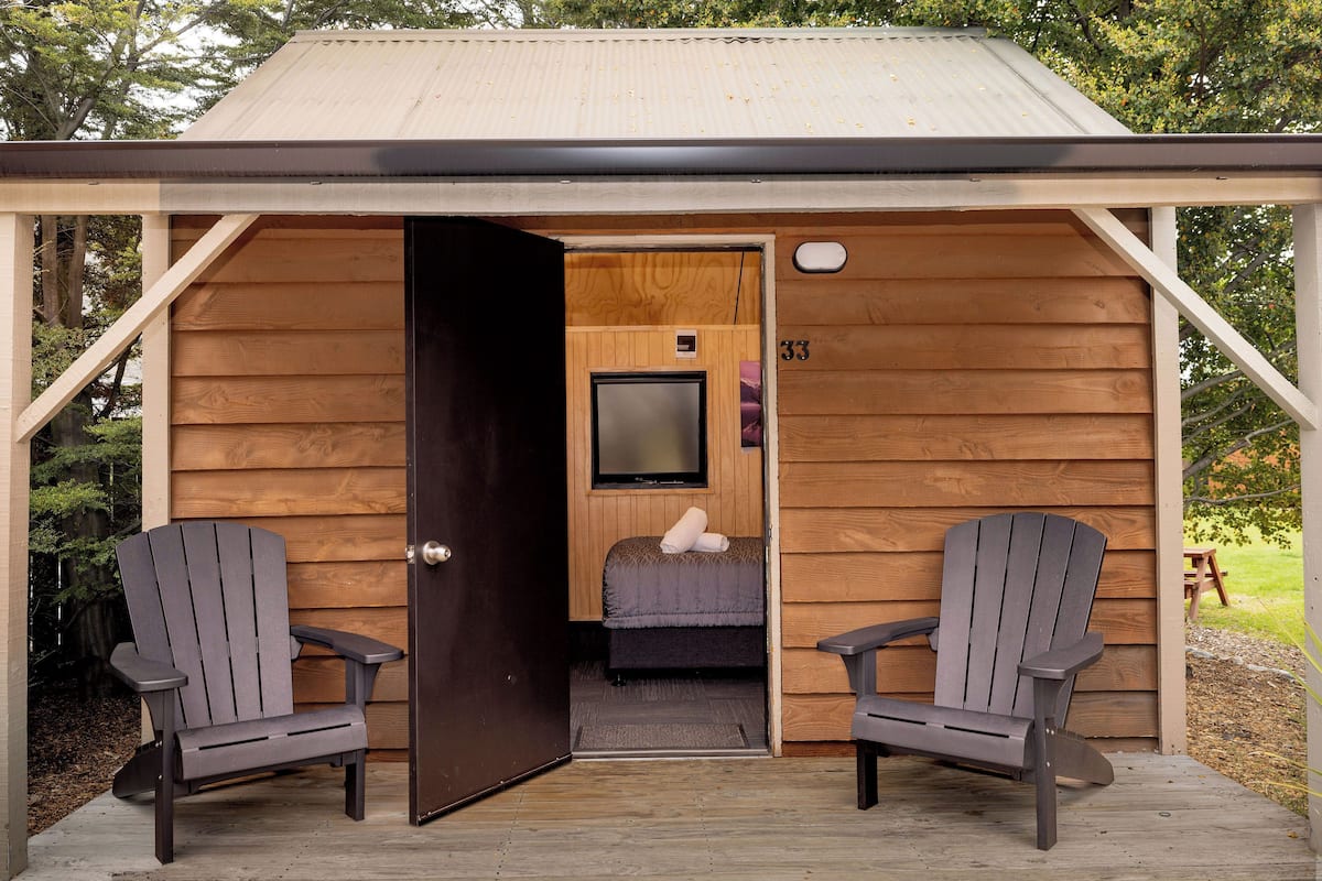 A wooden cabin with a door leading to a room containing a bed and a television. There are two Adirondack chairs outside.