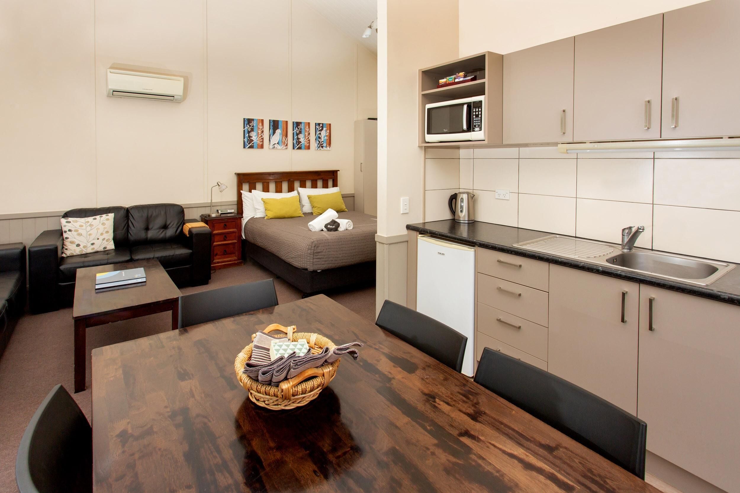 A compact living space with a sofa, a bed with pillows, a dining table with chairs, a kitchen area with a microwave, and a small kitchenette.