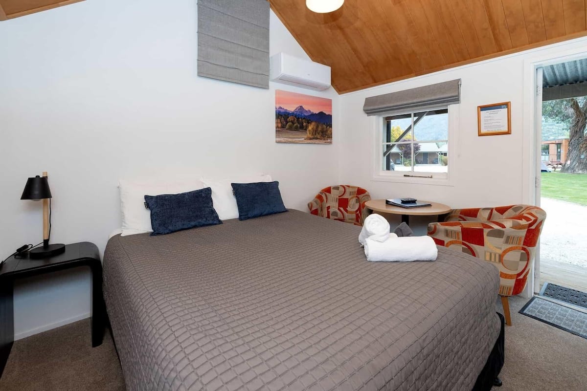 A bedroom with a large bed, a bedside table with a lamp, a chair, a small table, and a window with a view of the outdoors.
