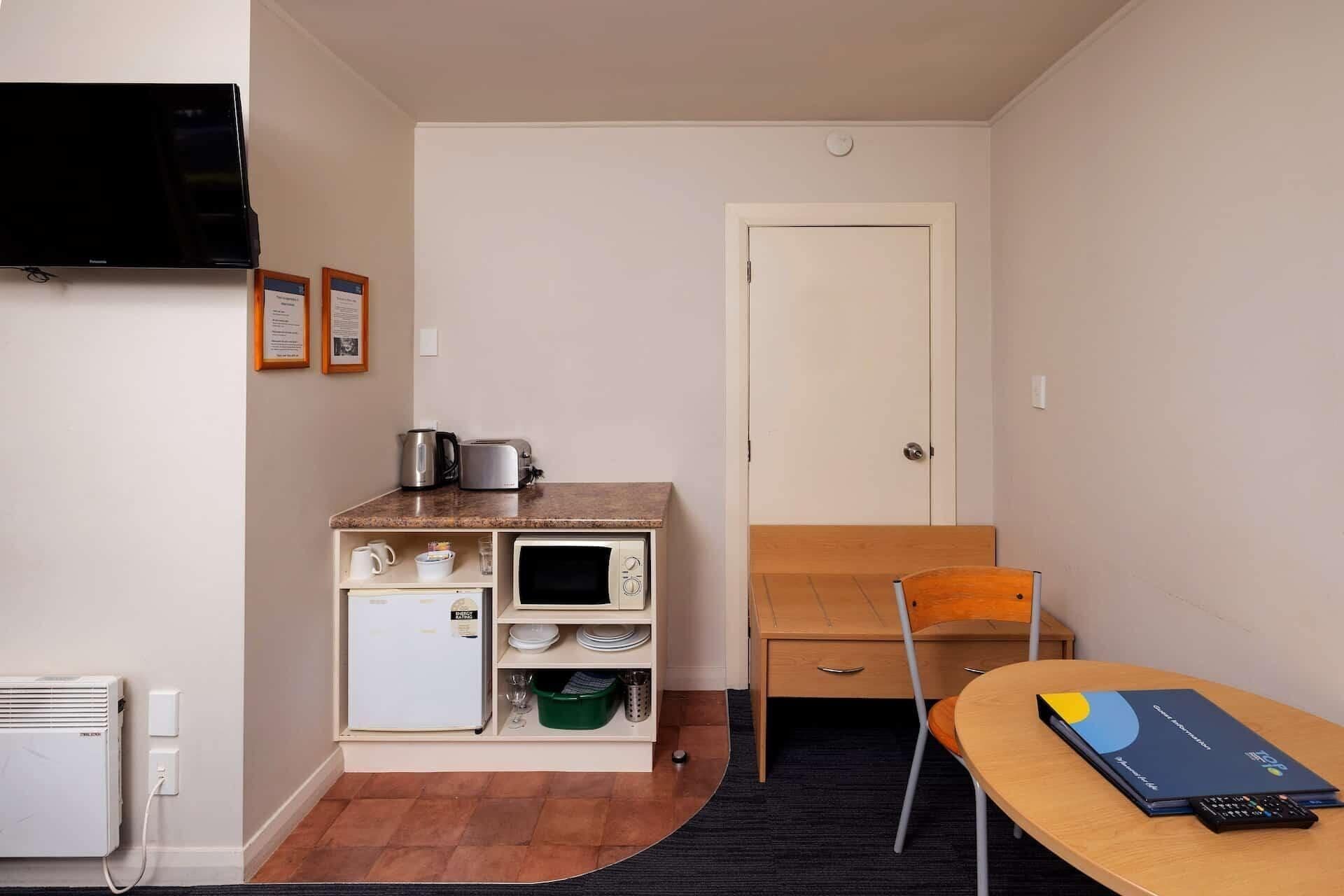 Une petite cuisine équipée d’un micro-ondes, d’un grille-pain et d’une bouilloire, d’un petit réfrigérateur, d’une chaise en bois et d’une table sur laquelle se trouvent un livre et une télécommande.