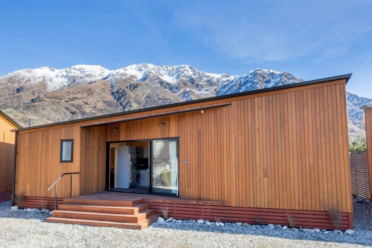 A modern wooden building with a staircase leading to a glass door entrance, set against a mountainous backdrop.