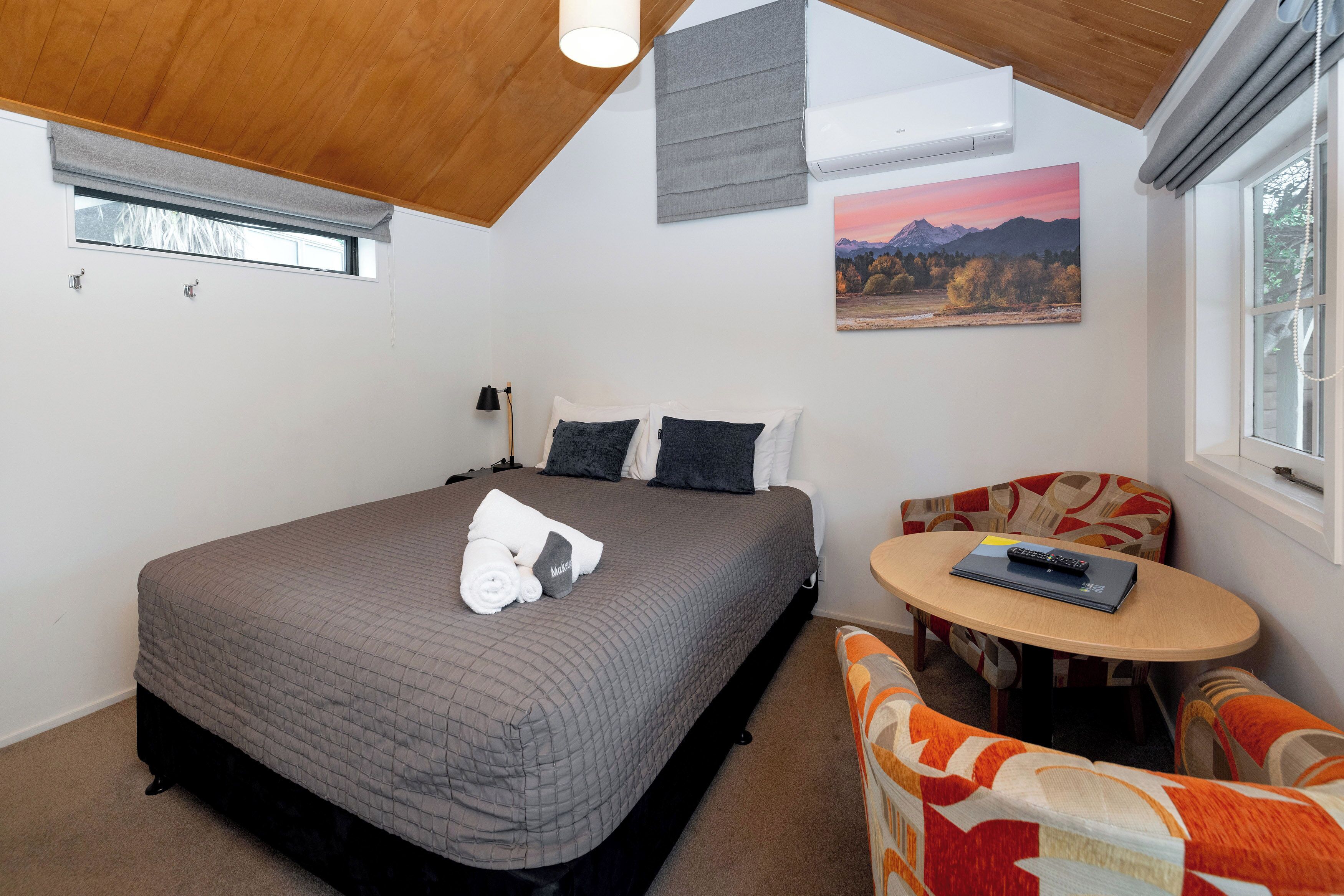 A bedroom with a bed, a small round table, and a chair. There is a window with a view of mountains and a painting on the wall.