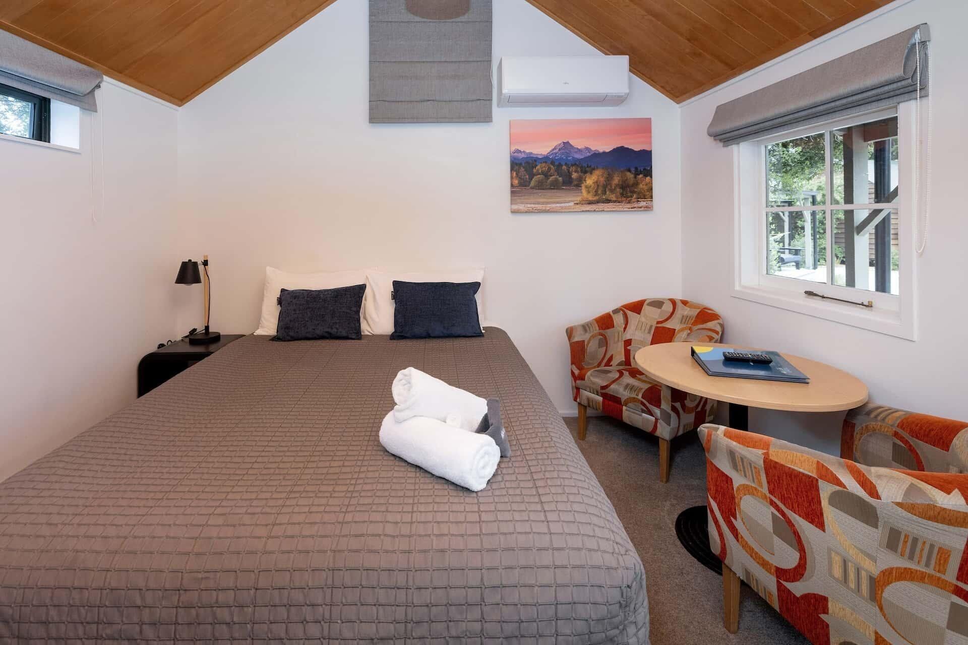 A bedroom with a large bed, a bedside table with a lamp, a chair, a small round table with a book and remote, and a window with a view of trees and mountains.