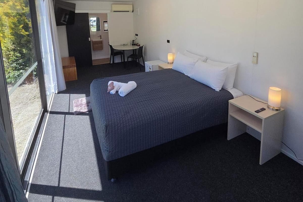 A modern hotel room with a large bed, bedside tables, and a view through a large window.