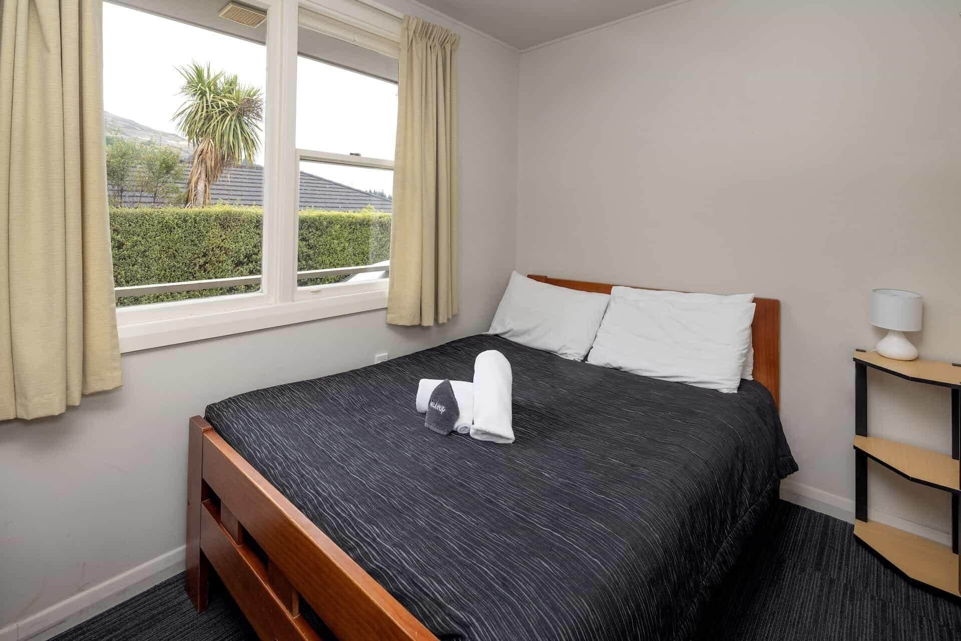 Hotelkamer met een bed, een raam met gordijnen, een nachtkastje met een lamp en uitzicht op groen.