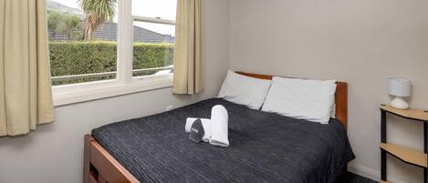 Una habitación de hotel con una cama, una ventana con cortinas, una mesita de noche con lámpara y vistas al exterior con vegetación.