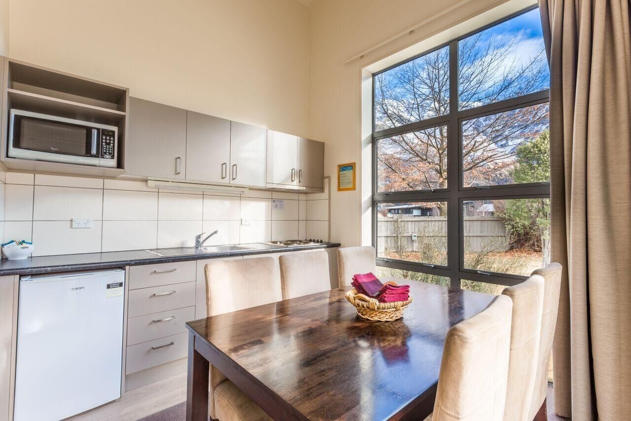 A modern kitchen with white cabinets, a microwave, and a small refrigerator. There is a dining table with chairs and a window with a view of trees.