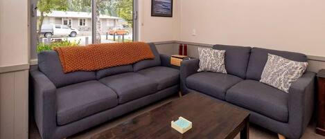 A living room with a dark brown coffee table, two grey sofas, and a window with a view of parked cars and greenery outside.