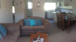 A living room with a brown sofa, a coffee table with a magazine and flowers, and a dining area with wooden chairs and a table. - The Peninsular Merimbula (Merimbula)