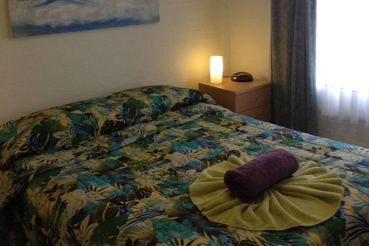 A hotel room with a bed, a painting of a beach scene, a ceiling fan, and a nightstand with a lamp.
