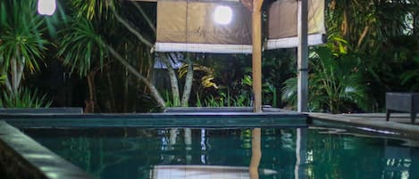 A swimming pool with a tiled deck, surrounded by tropical plants and a covered seating area.