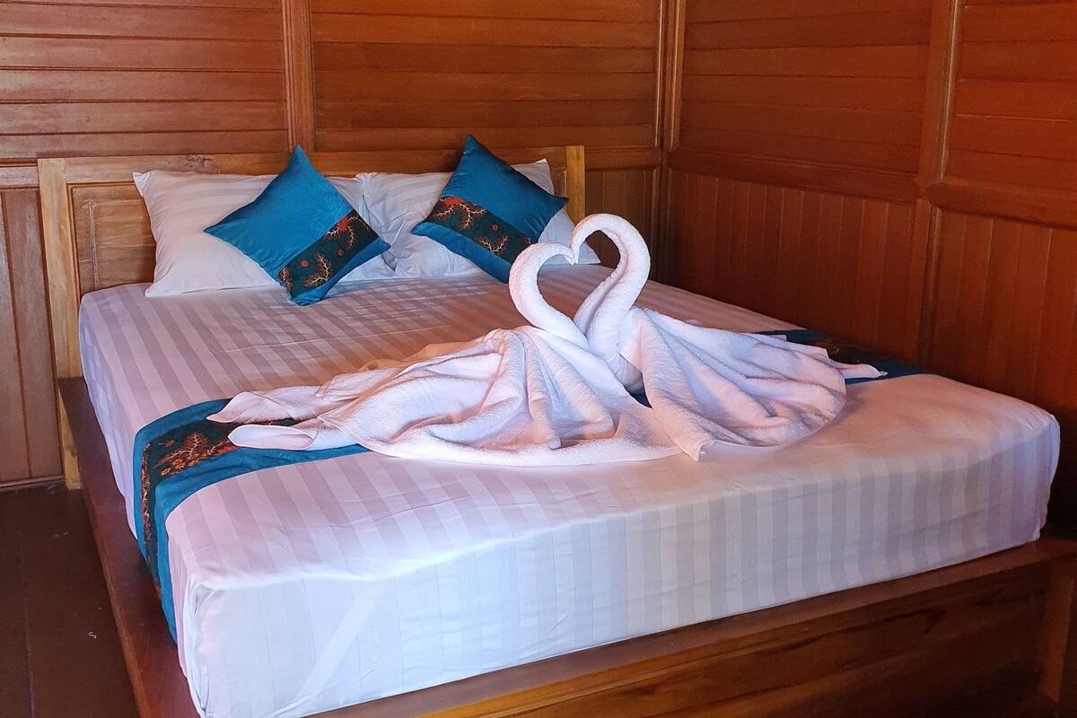 A wooden bed with a white mattress, blue pillows, and a swan-shaped towel arrangement.