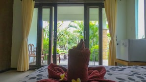 A hotel room with a bed covered in a red towel, a yellow flower, and a view of a garden through a sliding door. - Medori Putih Homestay (Pecatu)