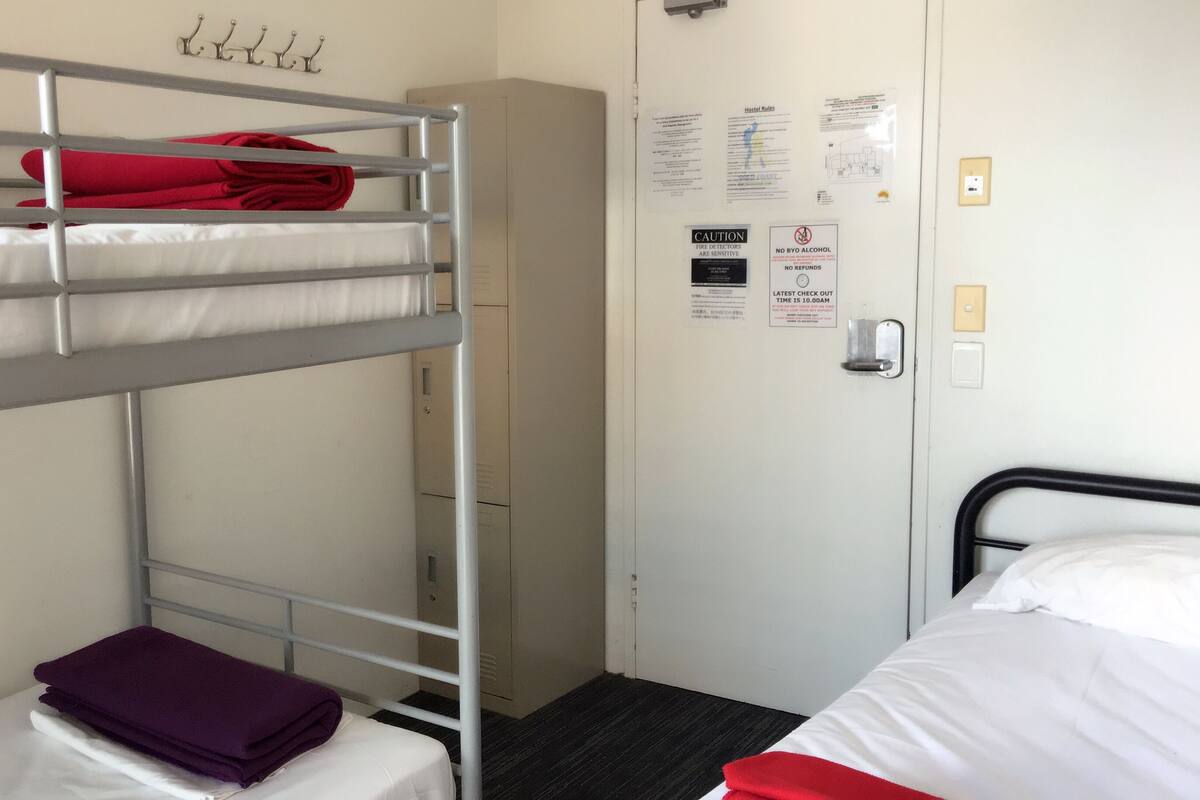 A bunk bed room with a red towel on the top bunk, a purple towel on the bottom bunk, and a white bed with a red blanket. There are metal hooks on the wall above the bunk bed and a door with various notices and a light switch.