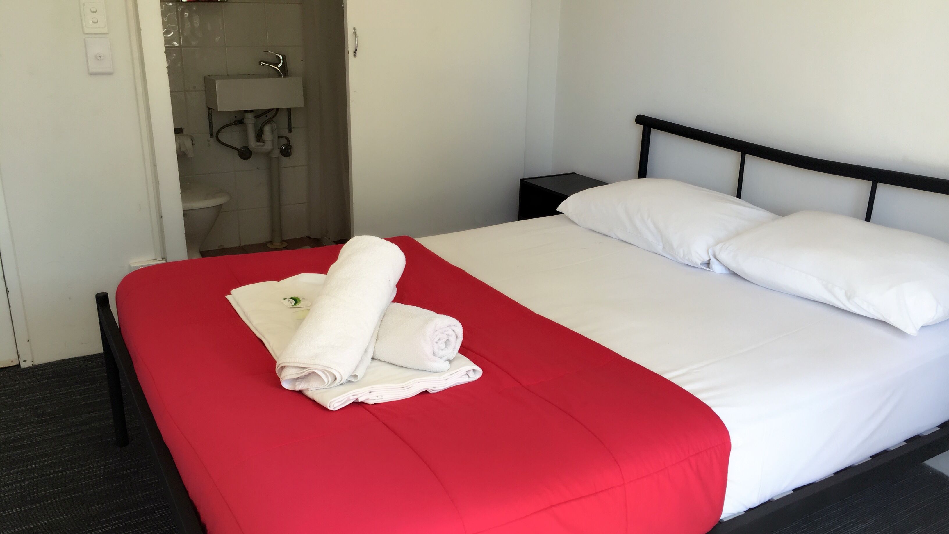Ett hotellrum med en snyggt bäddad säng, ett rött överkast, vita kuddar och en hopvikt handduk på sängen.