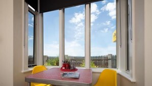 Une pièce avec une table et deux chaises jaunes, de grandes fenêtres donnant sur une clôture et le ciel.