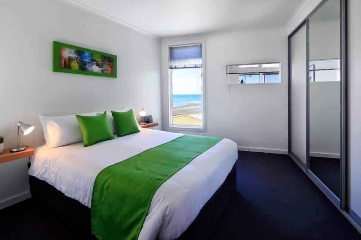 A hotel room with a bed, a nightstand, a lamp, a window with a view of the beach, and a sliding door.