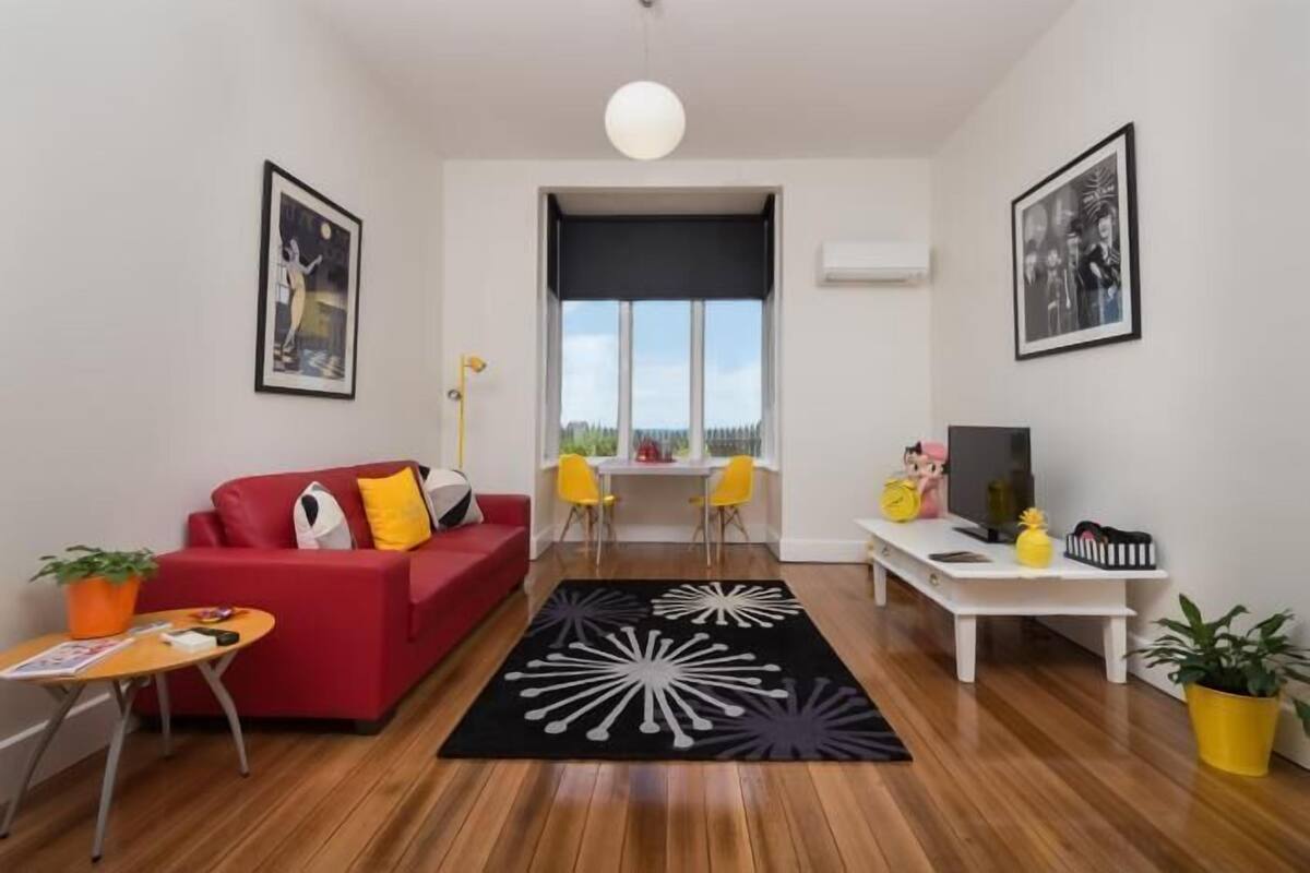 A modern living room with a red sofa, yellow chairs, a white coffee table, and a black and white rug. There is a TV on a white stand, and a window with a view of the outside.