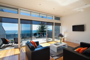 A modern living room with a large glass door leading to a balcony with a view of the ocean. There are two black sofas, a white coffee table, and a TV mounted on the wall. - Penguin Waterfront Escape (Penguin)
