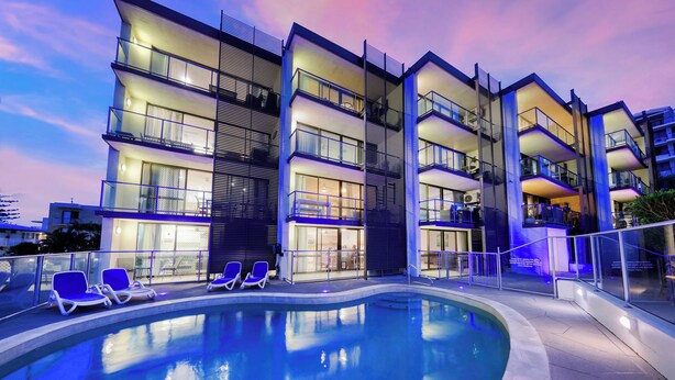 A modern hotel with a swimming pool, multiple balconies, and a well-lit exterior. - Merrima Court Holidays (Kings Beach)