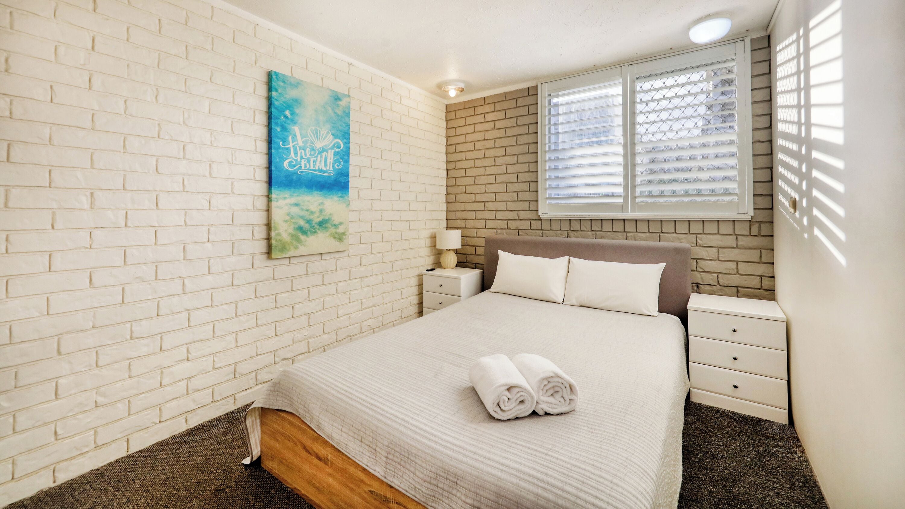 A bedroom with a white brick wall, a bed with a beige headboard, a window with blinds, a nightstand with a lamp, and a painting on the wall.