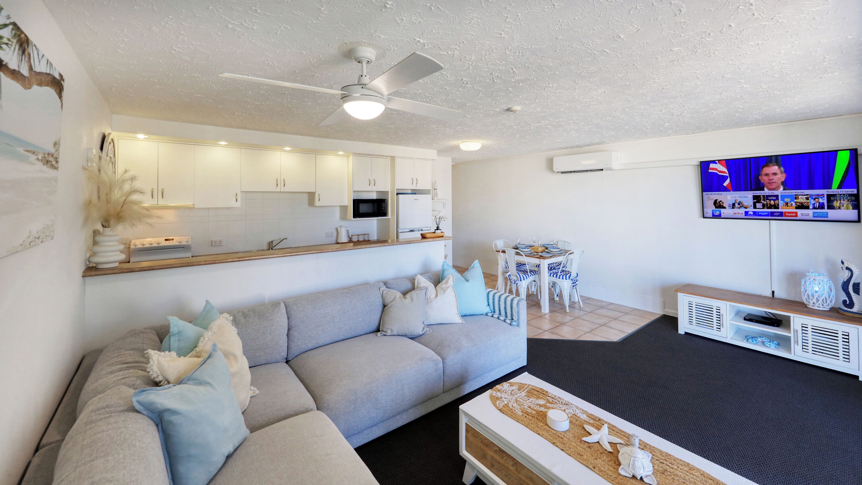 A modern living room with a grey sofa, a small side table, a flat-screen TV mounted on the wall, and a ceiling fan.