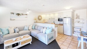 A modern living room with a grey sofa, a small coffee table, and a kitchen area with white cabinets and a microwave. - Merrima Court Holidays (Kings Beach)
