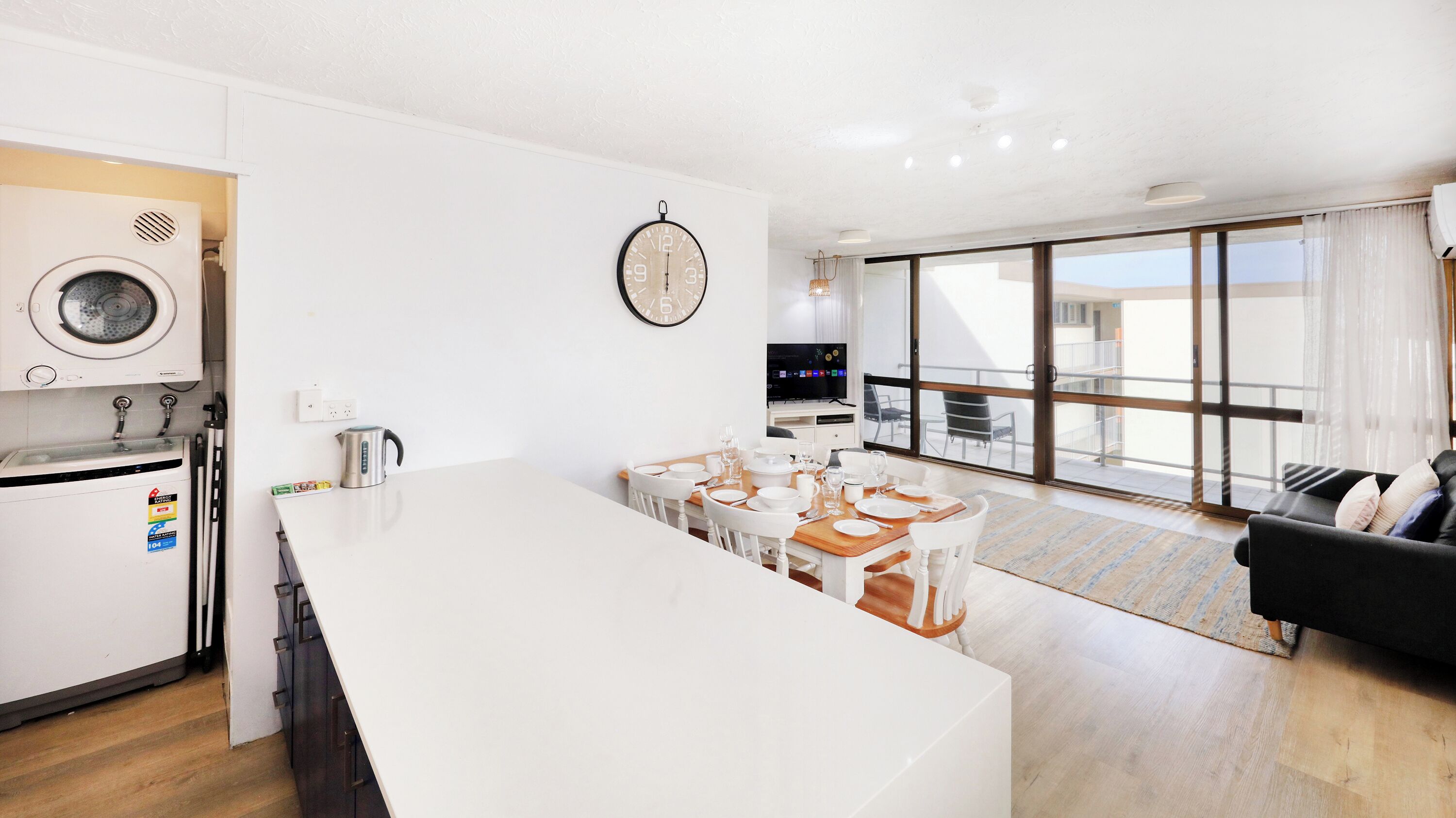 A modern kitchen with a built-in washer and dryer, a dining area with a wooden table and white chairs, a black sofa, and a large window offering a view of the city.