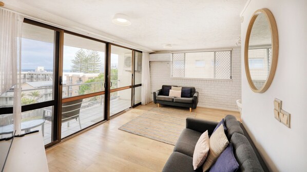 A spacious living room with a large sliding glass door leading to a balcony, a grey sofa with cushions, a small round table, and a view of buildings and trees.