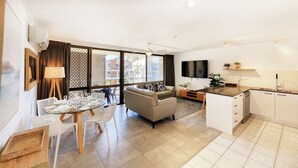 A modern living room with a sofa, a dining table set for four, and a kitchen area with a sink and countertop. - Merrima Court Holidays (Kings Beach)
