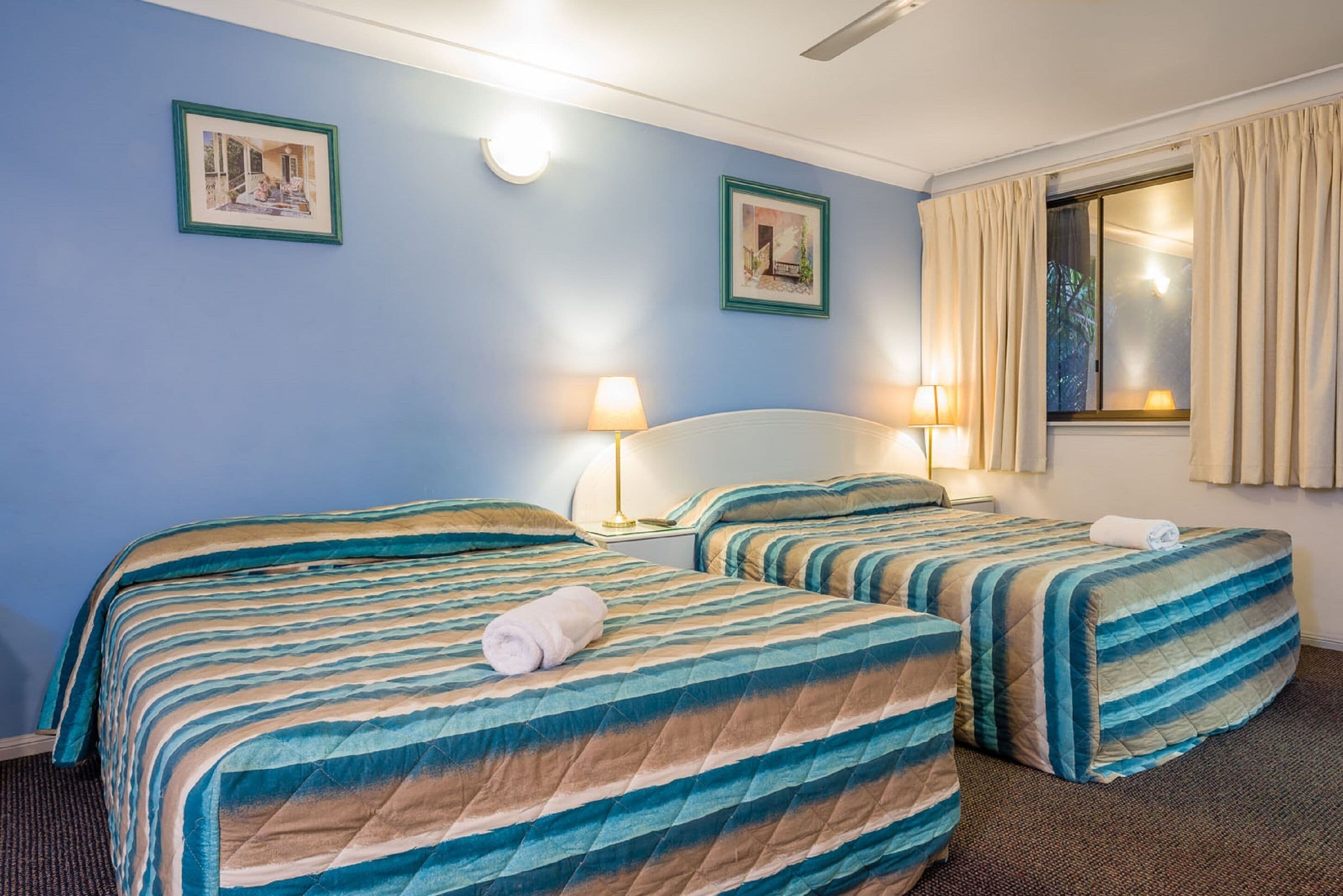 Een hotelkamer met twee bedden, elk voorzien van gestreept beddengoed, een nachtkastje met lampen en ingelijste foto's aan de muur.
