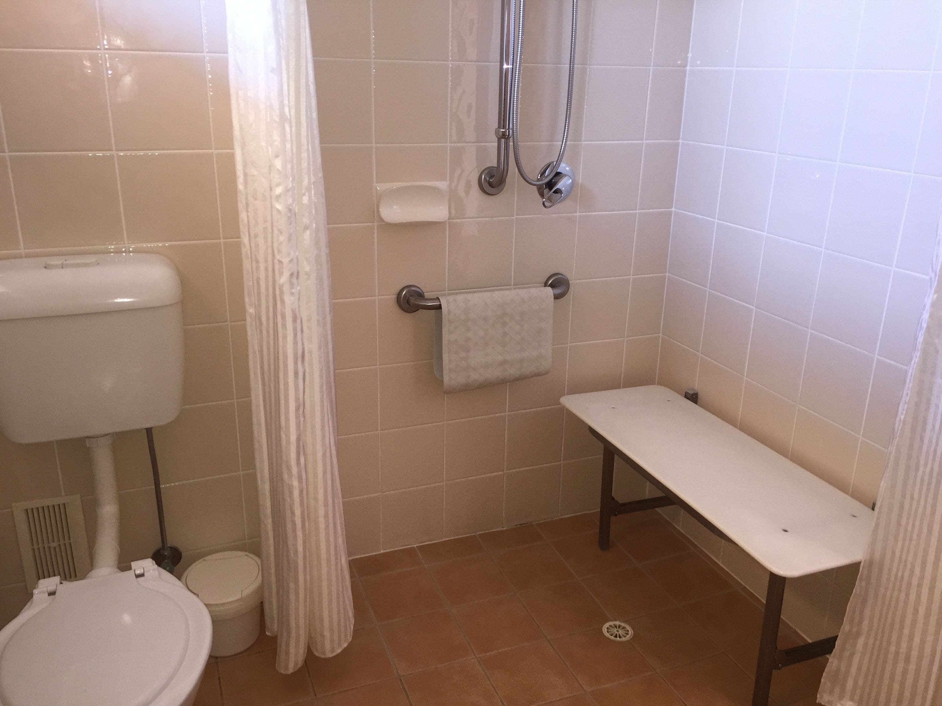A bathroom with a toilet, a shower with a handheld showerhead, a towel rack, and a bench.