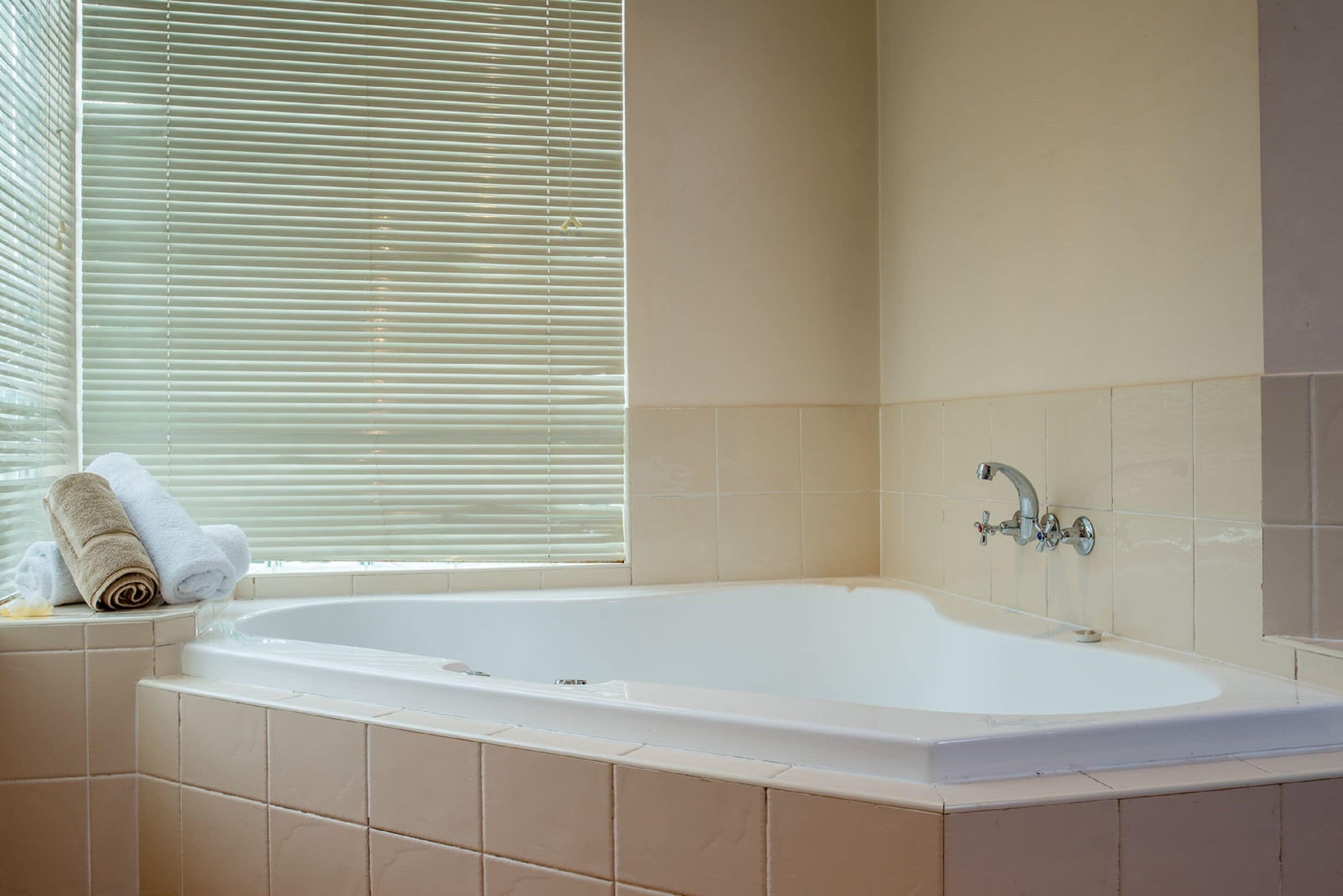 Une salle de bain avec une baignoire, des carreaux de couleur beige, une fenêtre avec des stores et un porte-serviettes avec des serviettes.