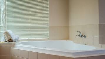 Une salle de bain avec une baignoire, des carreaux de couleur beige, une fenêtre avec des stores et un porte-serviettes avec des serviettes.