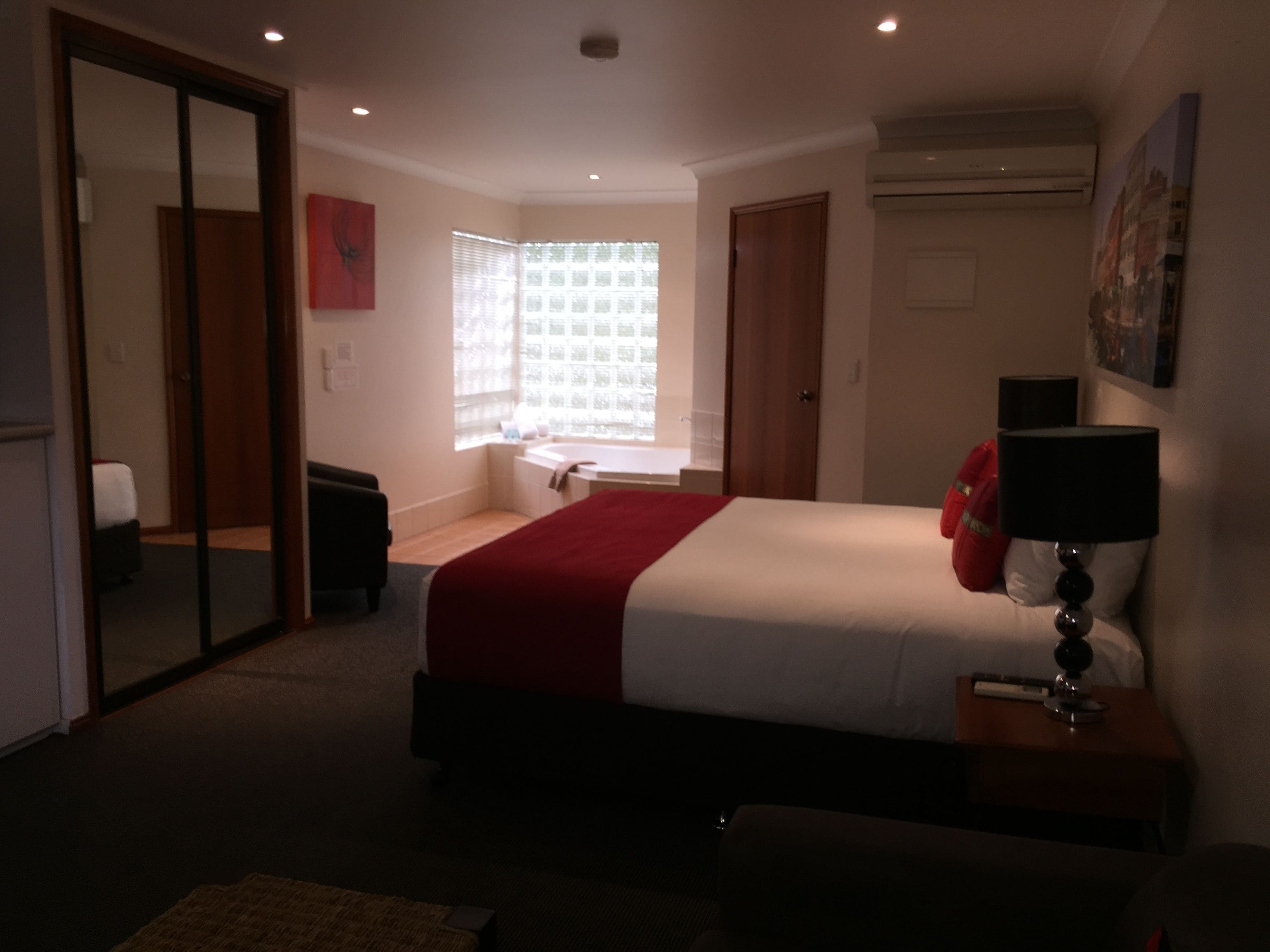 Una habitación de hotel con cama, ventana con persianas, mesita de noche con lámpara y una manta roja.