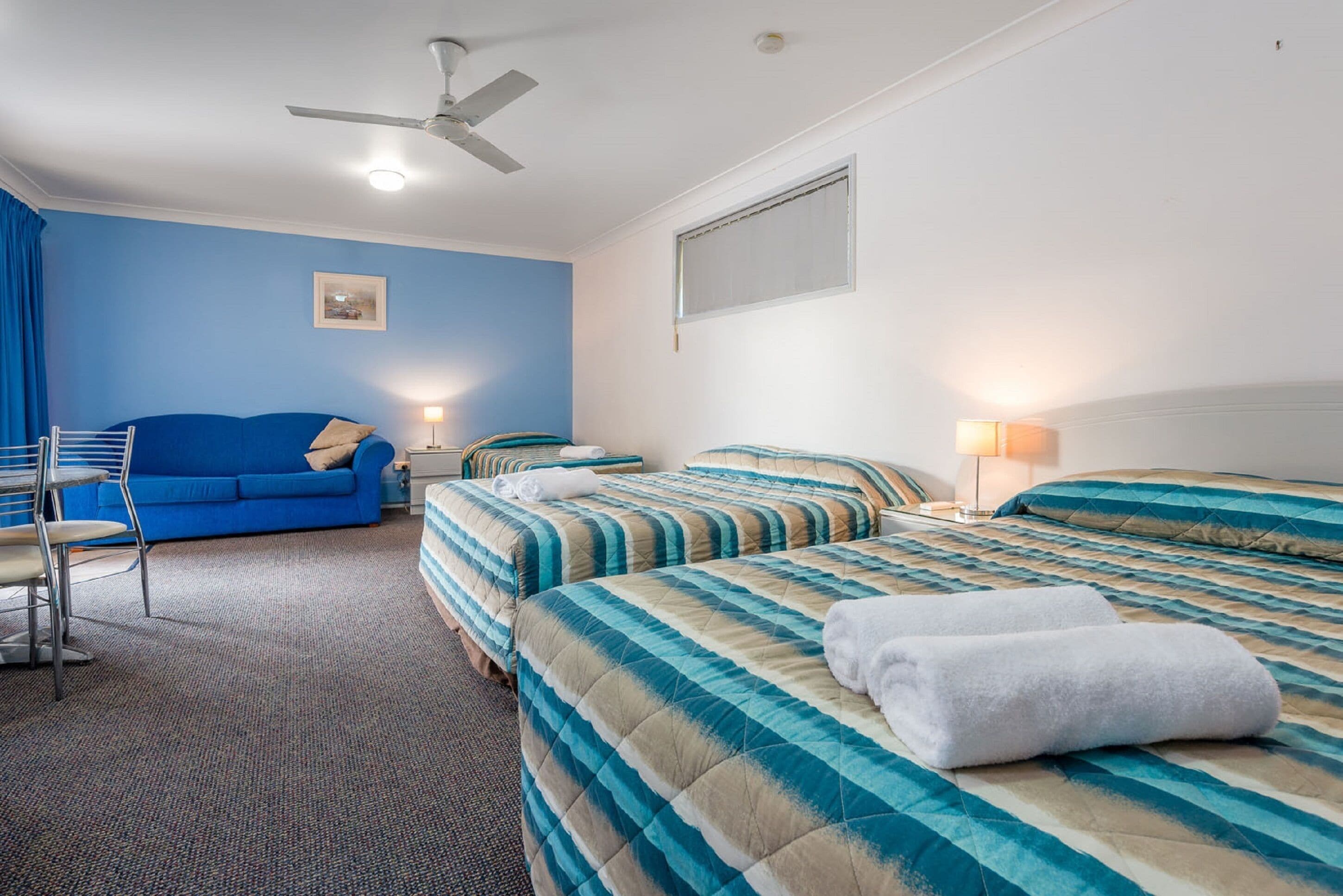A hotel room with two beds, a blue sofa, a ceiling fan, and a window with blinds.