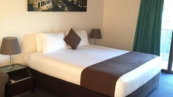A hotel room with a large bed, two bedside tables with lamps, a painting of a cityscape, an air conditioning unit, and a green curtain.